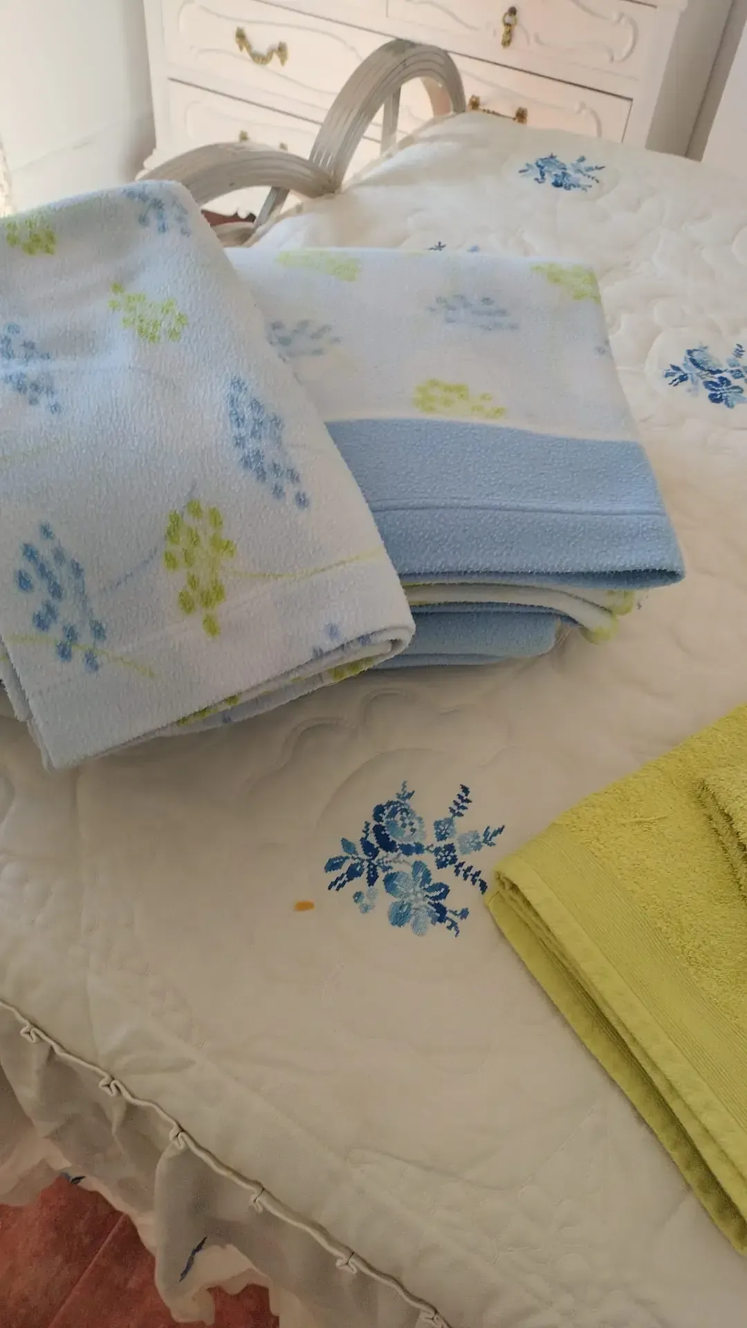 Toallas azules, blancas y amarillas dobladas sobre una cama con patrón floral bordado.