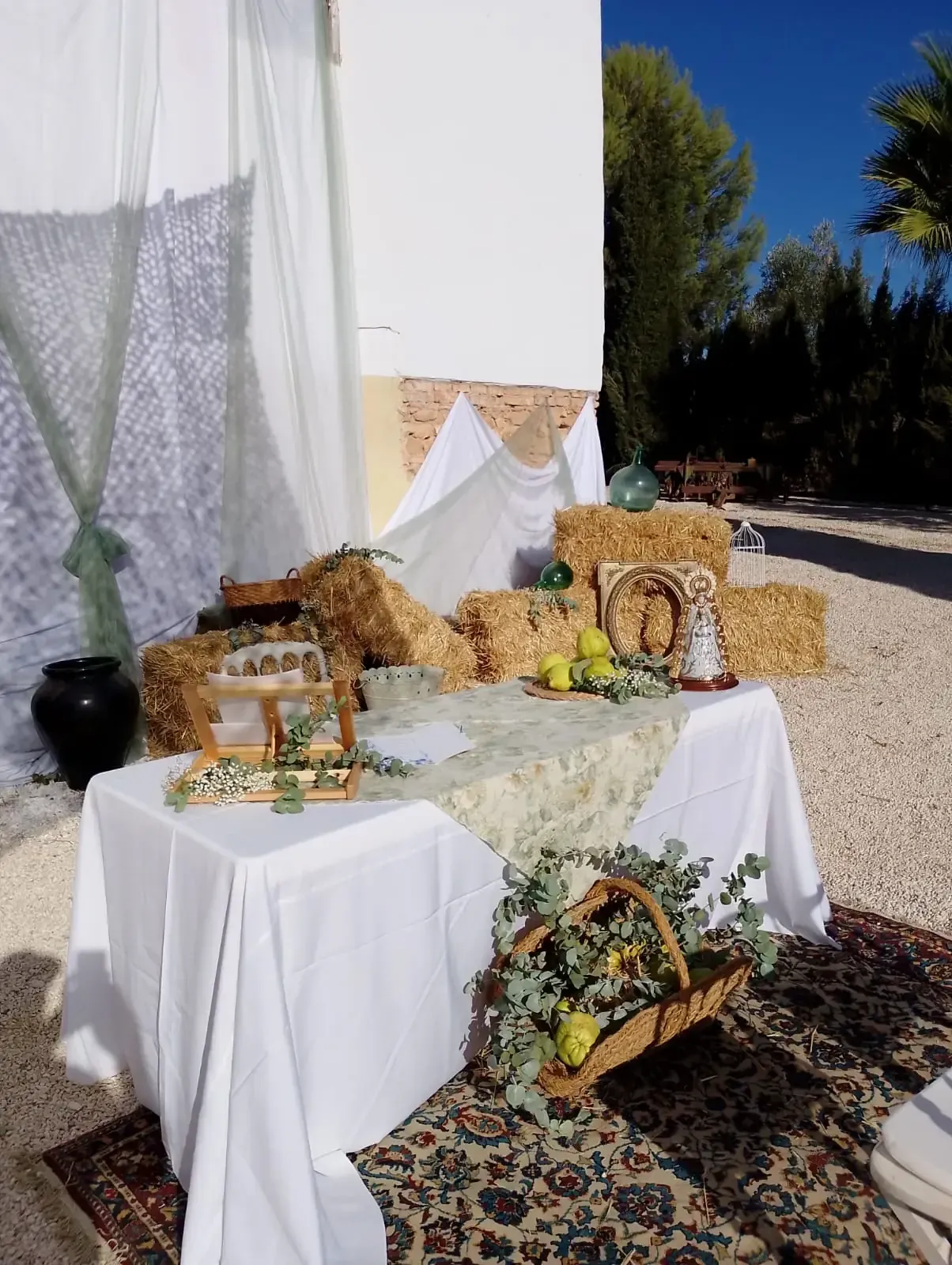 Exhibición al aire libre con fardos de heno, mesa con mantel blanco, arreglos florales y elementos decorativos.