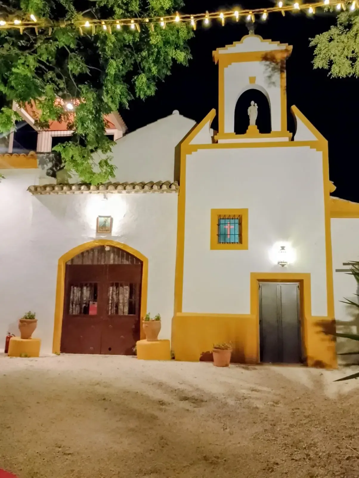 Edificio blanco con detalles amarillos, iluminado por la noche. Cuenta con un campanario, una puerta arqueada.