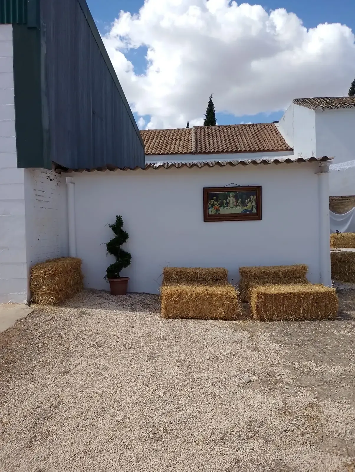 Pared blanca con arte enmarcado, topiario en espiral y asientos hechos con pacas de heno sobre una superficie de grava.