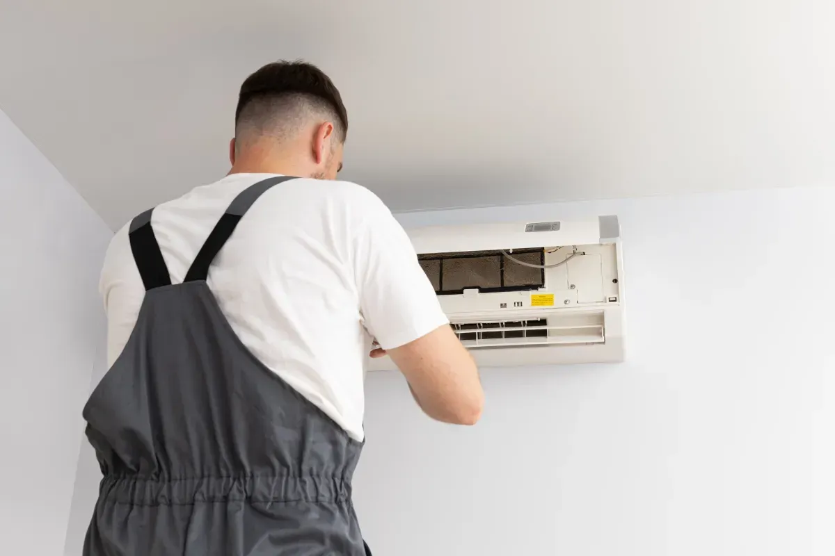 Una persona con un mono gris repara una unidad de aire acondicionado en una pared blanca.