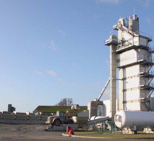Usine d'asphalte avec une haute tour, des réservoirs et une chargeuse par une journée ensoleillée.