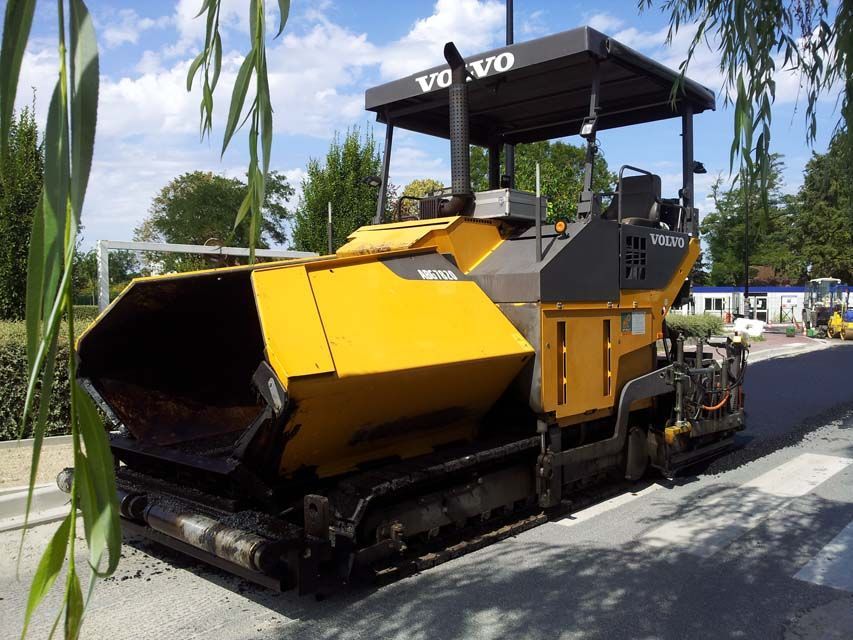 Finisseur d'asphalte Volvo jaune sur une route, sous un ciel bleu, près d'arbres.