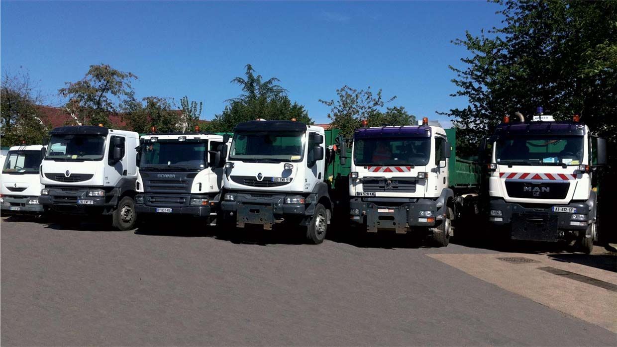 Six camions utilitaires blancs et bleus garés en rang sur l'asphalte, sur fond de ciel bleu.