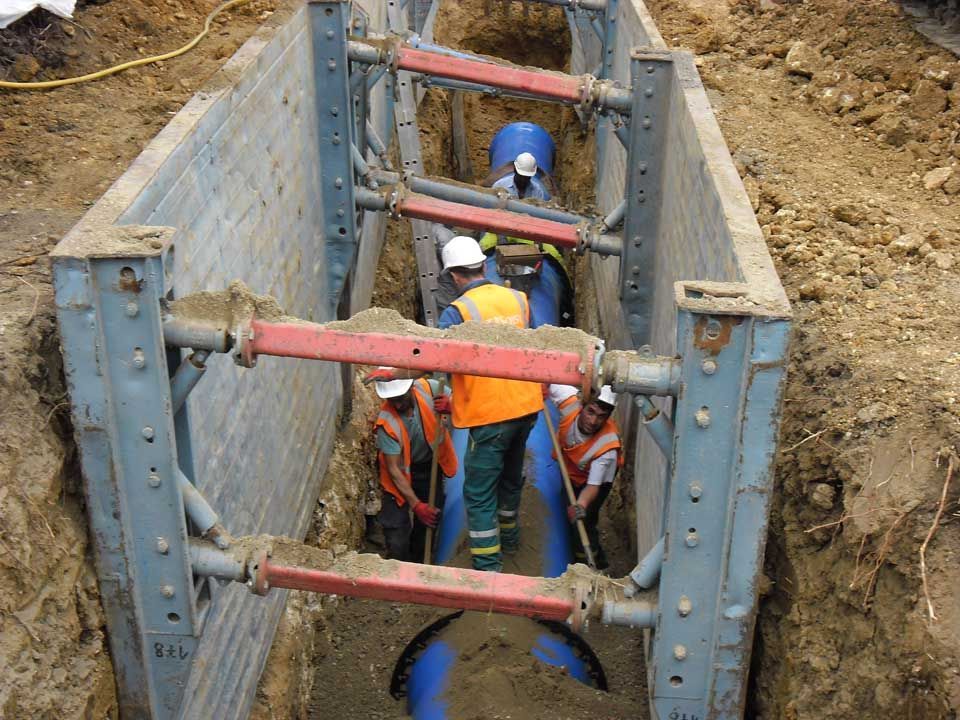 Des ouvriers installent un tuyau bleu dans une tranchée. Des étaiements protègent l'excavation.