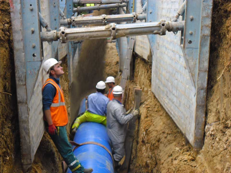 Des ouvriers du bâtiment installent un tuyau bleu dans une tranchée, soutenu par un étaiement en acier.