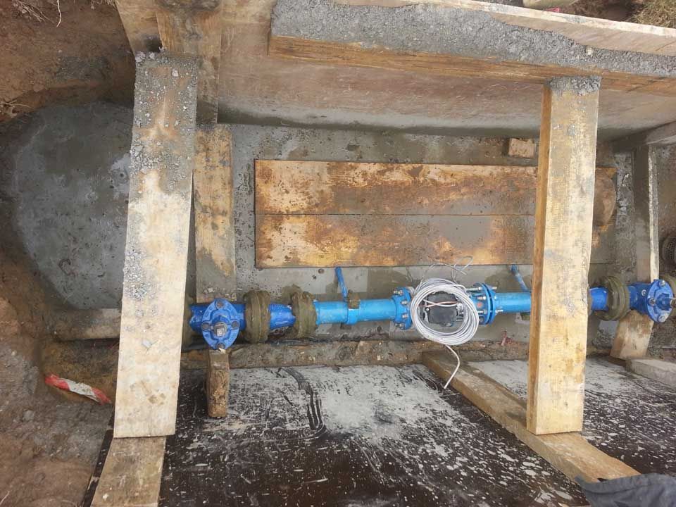 Tuyaux d'eau bleus avec vannes et compteur, dans une structure en bois à l'intérieur d'une enceinte en béton.