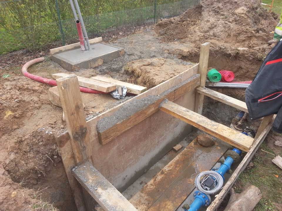 Chantier : Ossature en bois autour de tuyaux bleus dans une tranchée. Câbles rouges et verts visibles.