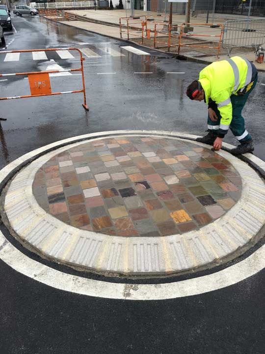Une personne portant un gilet fluo inspecte un petit rond-point carrelé sous la pluie.