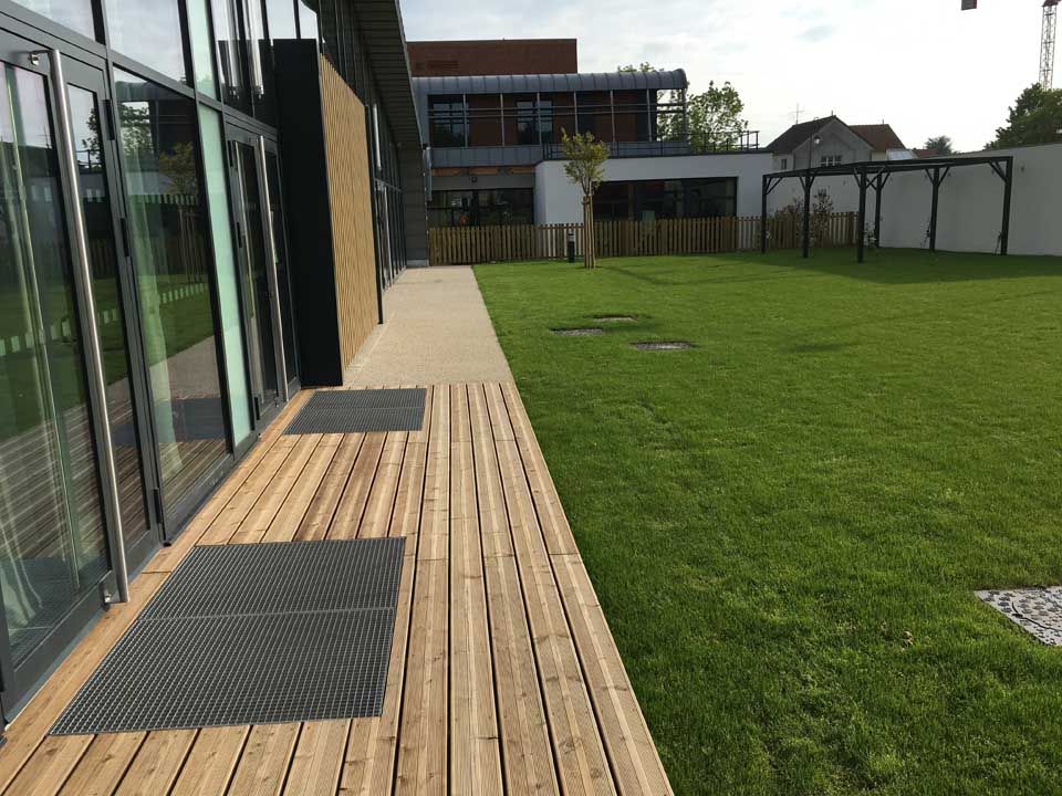 Terrasse en bois et pelouse jouxtant un bâtiment moderne doté de portes et fenêtres vitrées.