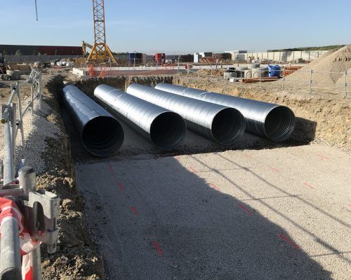 Quatre gros tuyaux métalliques ondulés posés dans une tranchée sur un chantier de construction.