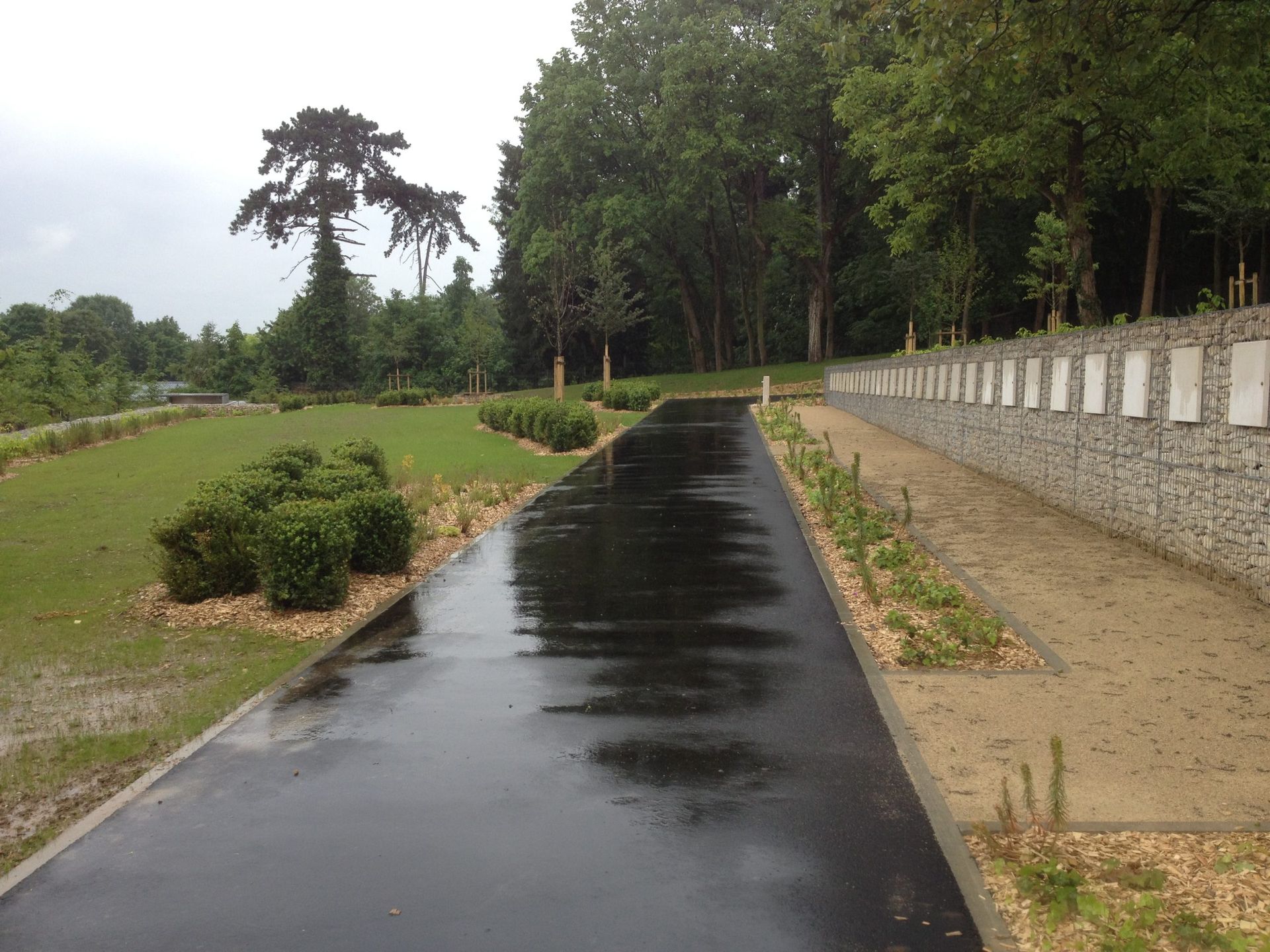 Chemin asphalté mouillé dans un parc paysagé, avec un mur de pierre orné de plaques et des arbres.