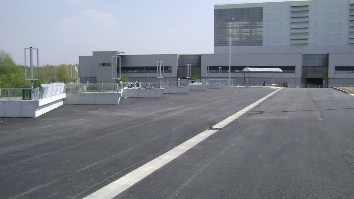 Parking asphalté désert devant un bâtiment gris moderne. Ciel bleu au-dessus.