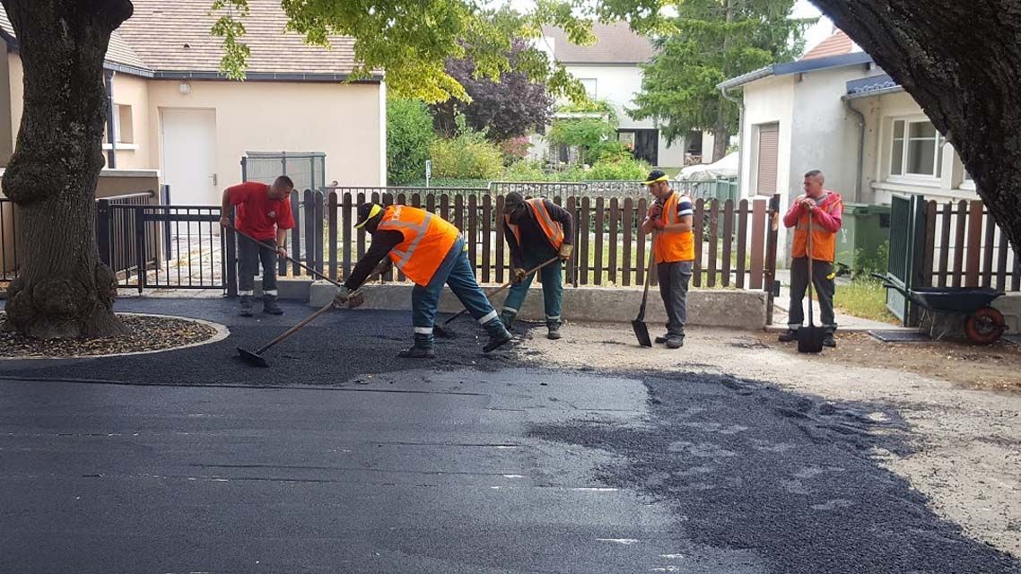Des ouvriers asphaltent une rue résidentielle ; tous portent des gilets orange, sous un arbre.