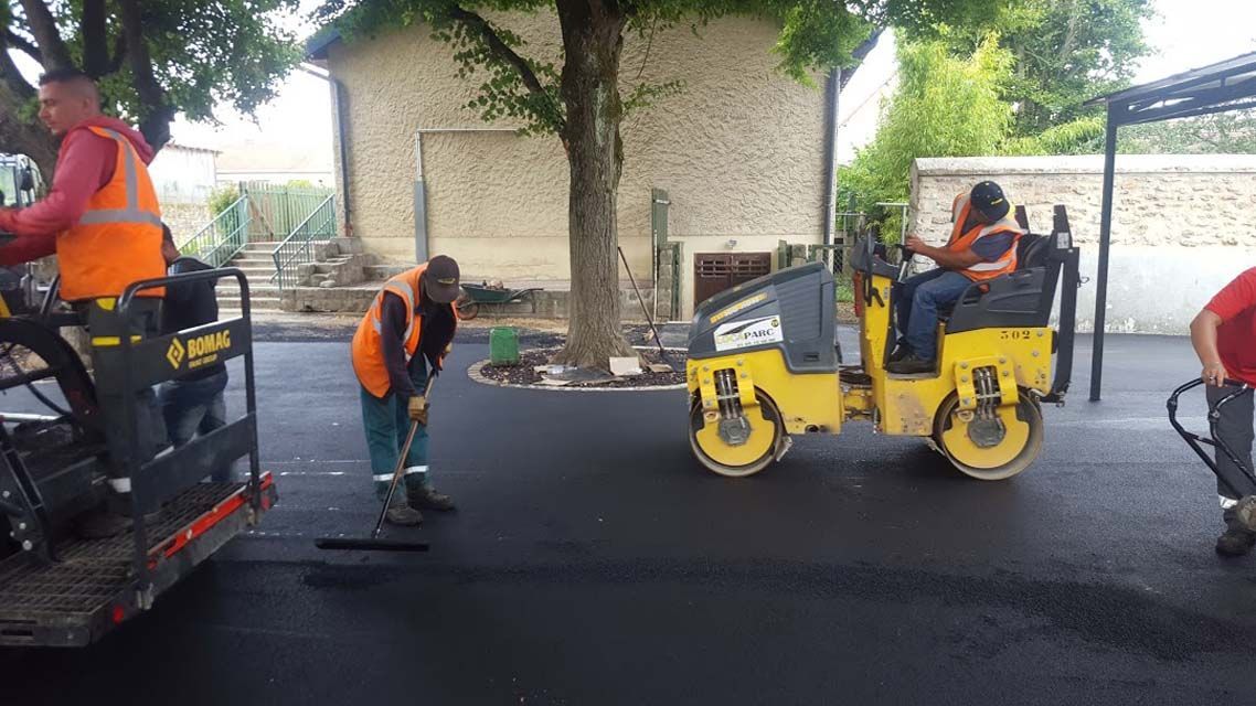 Travaux de voirie : Des ouvriers posent de l’asphalte, à l’aide d’un rouleau compresseur et d’une machine à paver, sur une rue, devant un bâtiment.