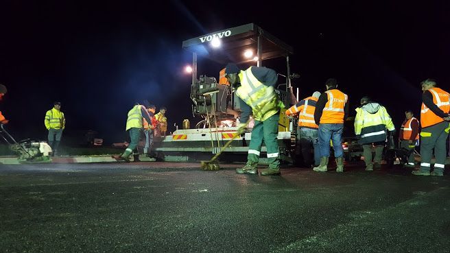 Une équipe de travaux routiers pose de l'asphalte de nuit ; les ouvriers portent des gilets réfléchissants près de la machine à asphalter sous un ciel sombre.