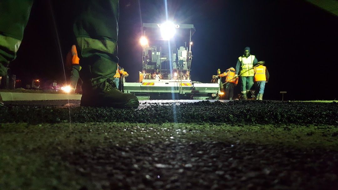Travaux de pavage d'asphalte de nuit. Des ouvriers en gilets de sécurité travaillent autour d'une machine à paver sous des projecteurs puissants.