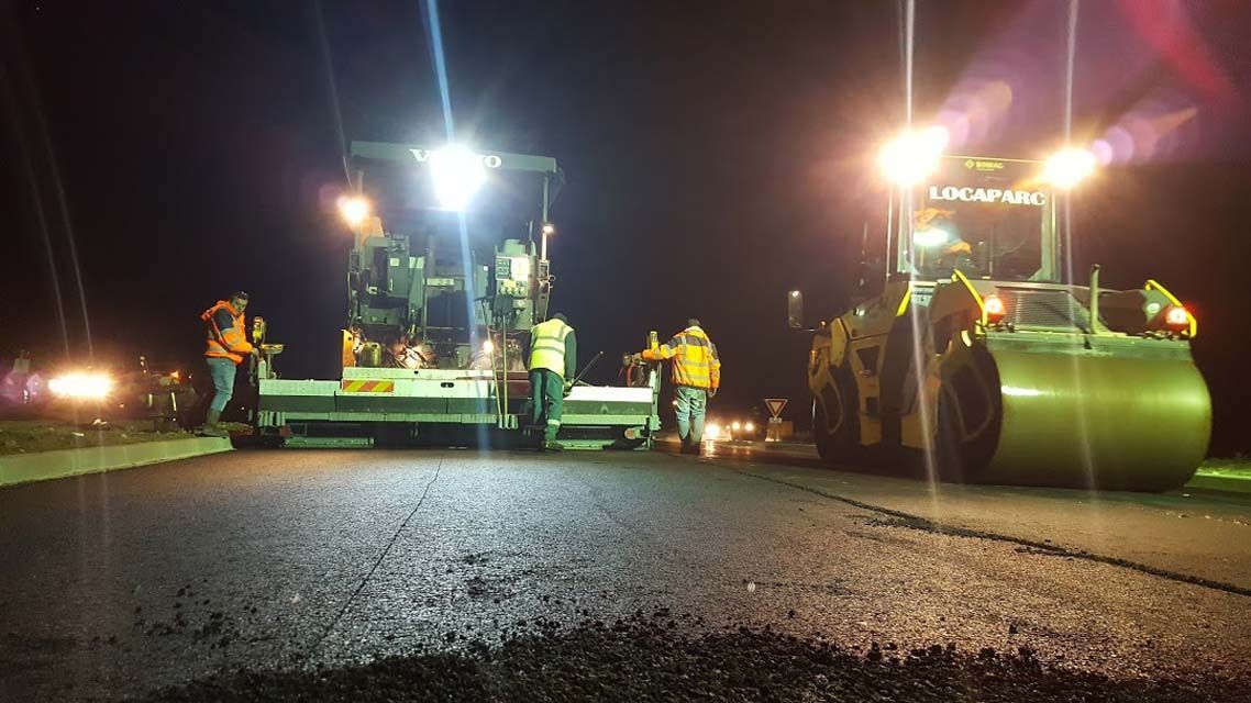 Travaux de pavage nocturnes ; ouvriers, finisseur et rouleau compresseur sous des projecteurs. Pose d’asphalte.