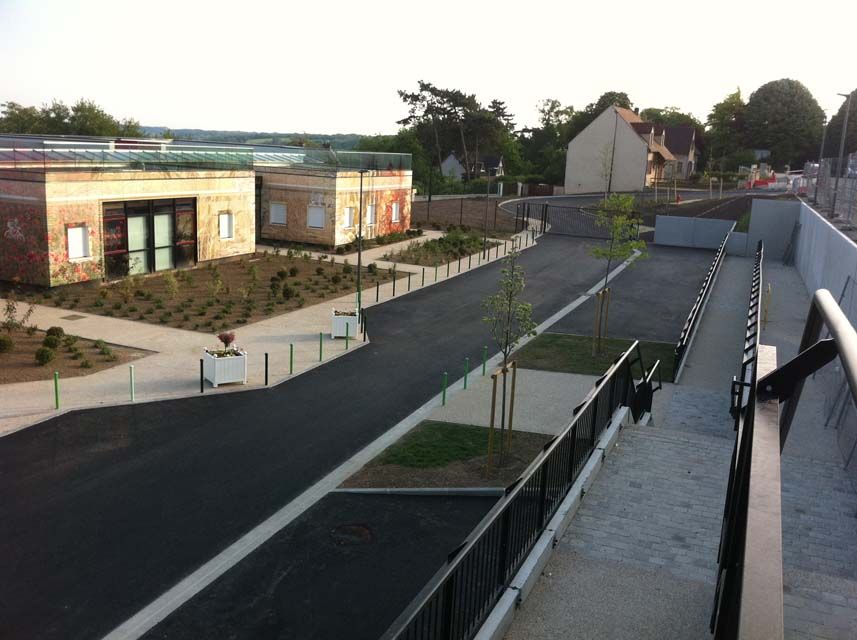 Rampe d'accès en asphalte et chemin agrémenté de plantations, menant à des bâtiments et des habitations baignés de lumière naturelle.