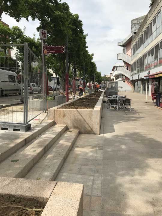 Travaux de construction sur le trottoir : marches, jardinières, clôture, ouvriers, panneau de métro et un bâtiment par une journée ensoleillée.