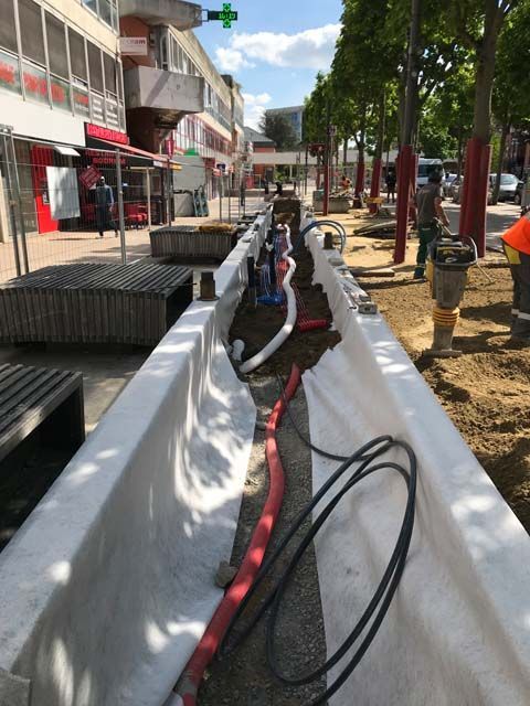 Chantier en pleine rue. Une longue tranchée à ciel ouvert, tapissée de tissu blanc, contient des tuyaux et des câbles.