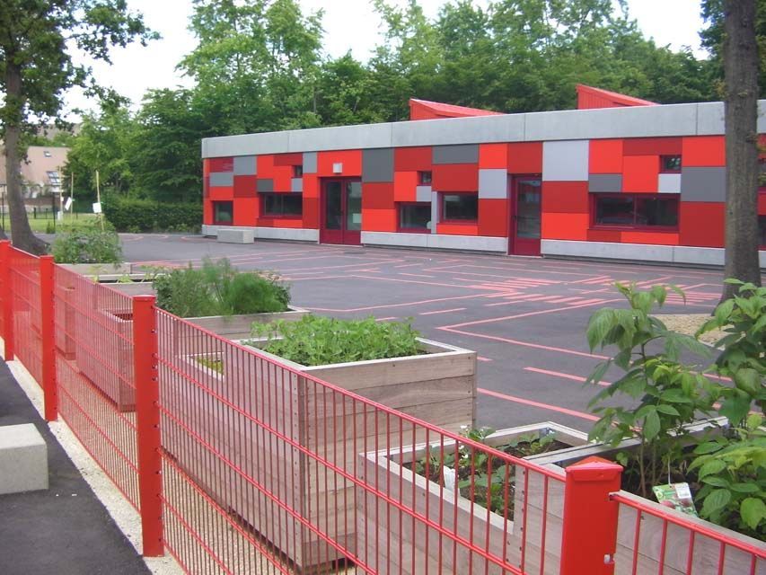 Bâtiment scolaire avec une façade rouge, grise et orange, une clôture rouge et des jardinières surélevées.