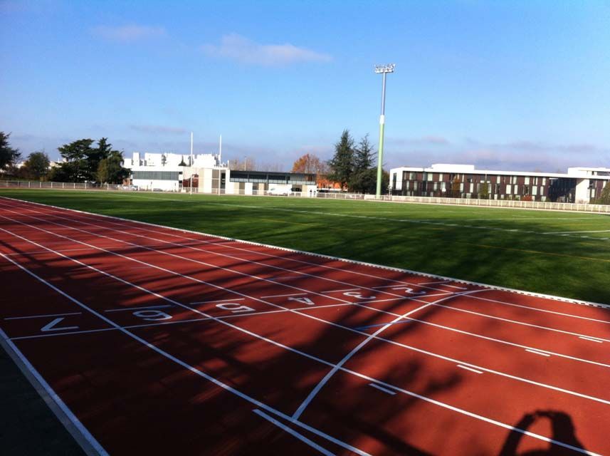 Piste d'athlétisme rouge avec marquages ​​au sol, pelouse verte et bâtiments modernes sous un ciel bleu.
