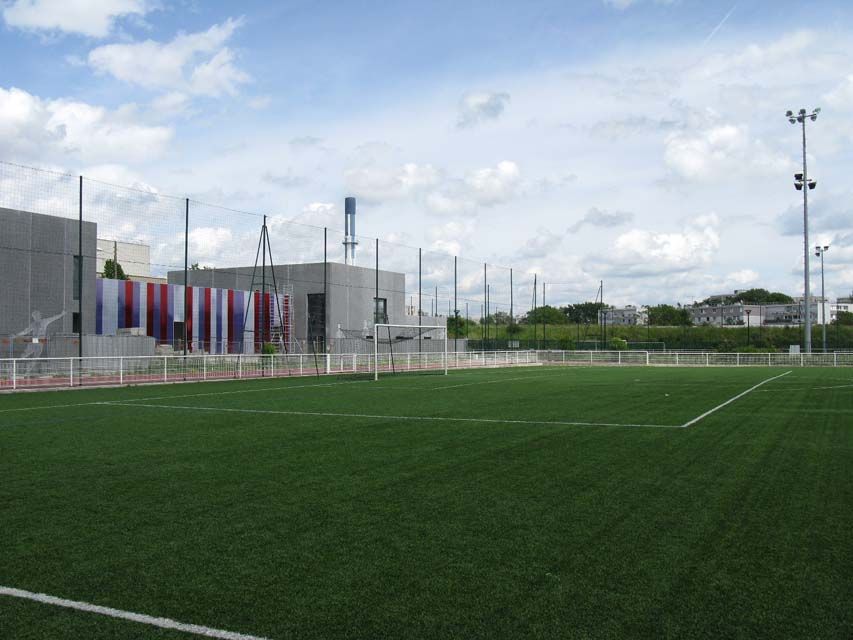 Terrain de football en gazon synthétique vert, bâtiment gris, ciel bleu, nuages ​​blancs.