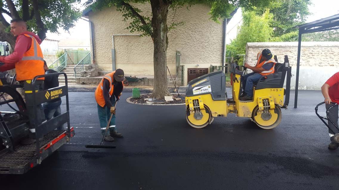 Une équipe de voirie effectue des travaux de pavage. Des ouvriers en gilets orange utilisent des engins, des pelles et un rouleau compresseur.