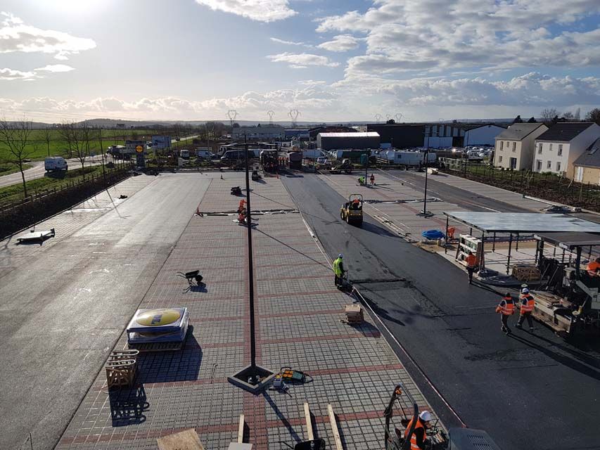 Des ouvriers asphaltent un grand parking ; journée ensoleillée.