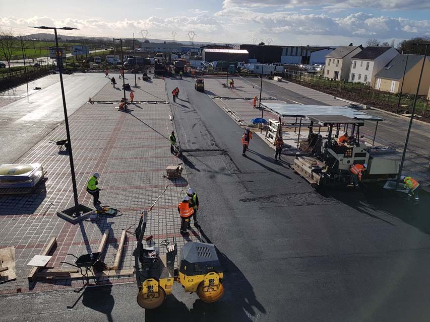 Des ouvriers du bâtiment asphaltent un parking. Pose d'asphalte, plusieurs ouvriers avec du matériel. Journée ensoleillée, quartier résidentiel en arrière-plan.