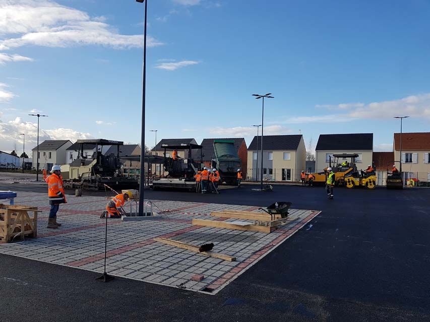 Chantier de construction avec des ouvriers posant des pavés près de maisons et de machines, sous un ciel bleu.