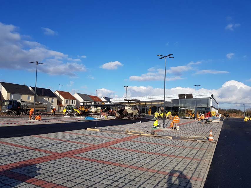Chantier de construction : ouvriers en gilets de sécurité posent des pavés, bâtiment commercial en arrière-plan, ciel bleu.