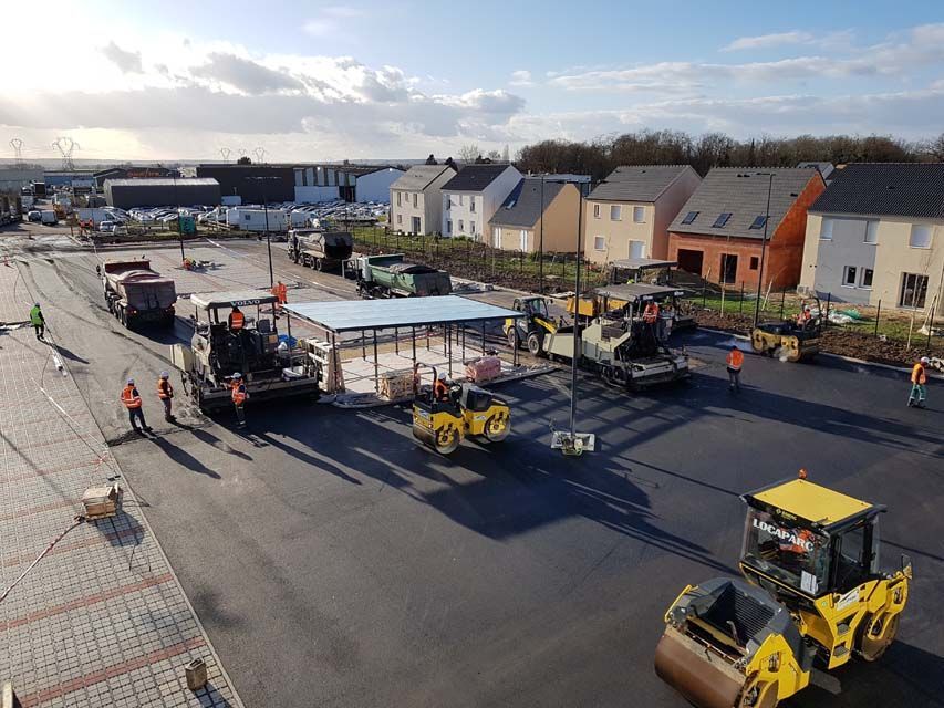 Une équipe de construction asphalte une route ; une zone résidentielle se trouve en arrière-plan.