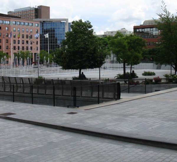 Place urbaine avec bâtiments, arbres et point d'eau. Pavés gris, architecture moderne et ciel nuageux.