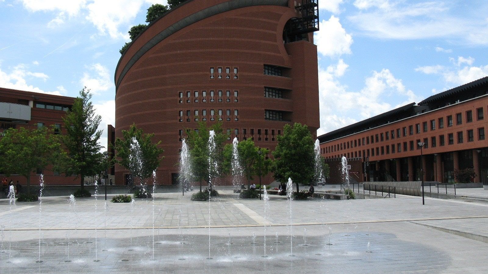 Place avec fontaine, grand bâtiment en briques et arbres sous un ciel bleu.