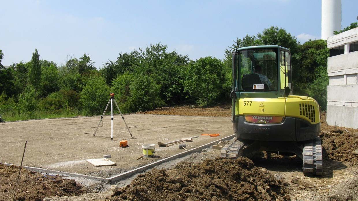 Une pelleteuse jaune sur un chantier de construction ; des arbres et un bâtiment blanc en arrière-plan.