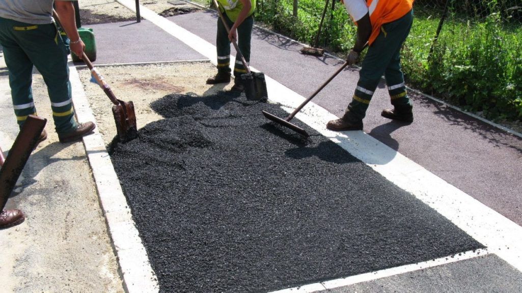 Des ouvriers routiers, vêtus de tenues de travail vertes et orange, répandent de l'asphalte sur une portion rectangulaire de chaussée.