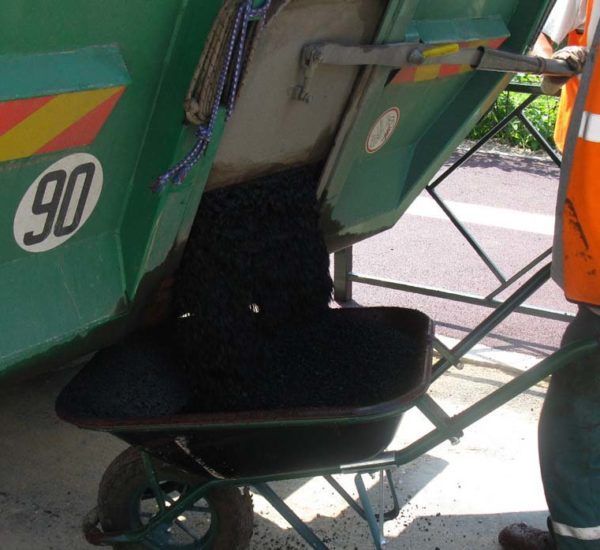 Une brouette remplie d'asphalte noir provenant d'un camion-benne vert, à côté d'une personne portant un gilet de sécurité.