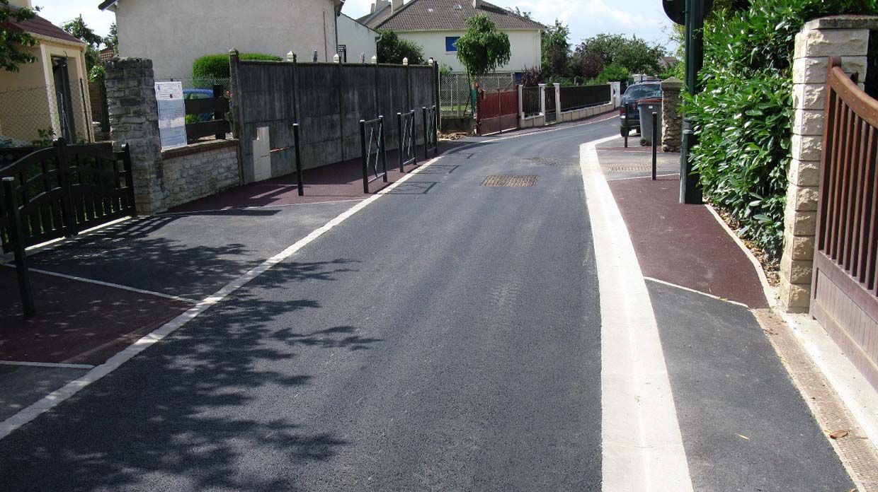 Route asphaltée avec ligne blanche, bordée de briques rouges et d'une clôture, bordée de bâtiments et de verdure.
