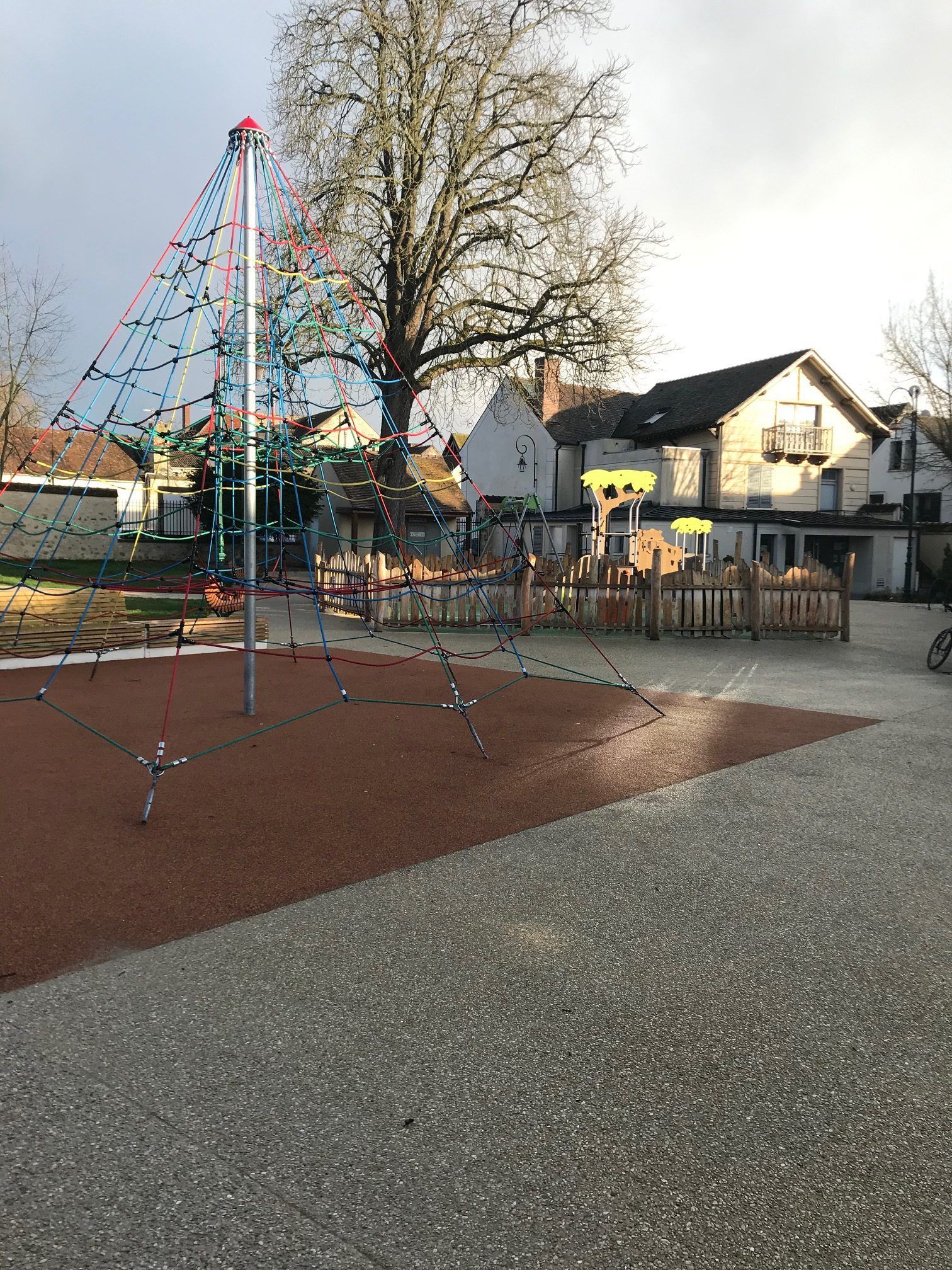 Aire de jeux avec structure d'escalade et clôture basse en bois, bâtiments et arbres en arrière-plan.