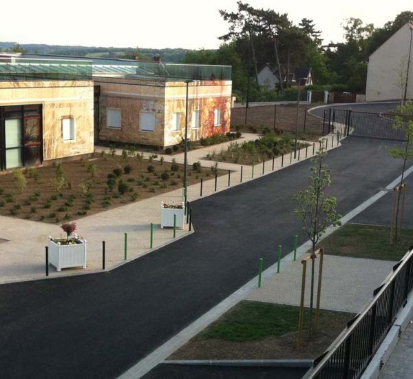 La route goudronnée serpente entre les bâtiments et les espaces verts. Des bornes vertes bordent la chaussée.