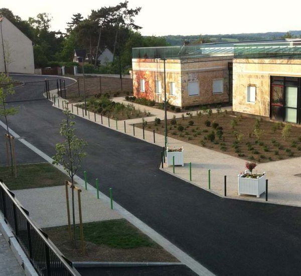 Route asphaltée bordée de verdure, de bâtiments et de jardinières blanches.