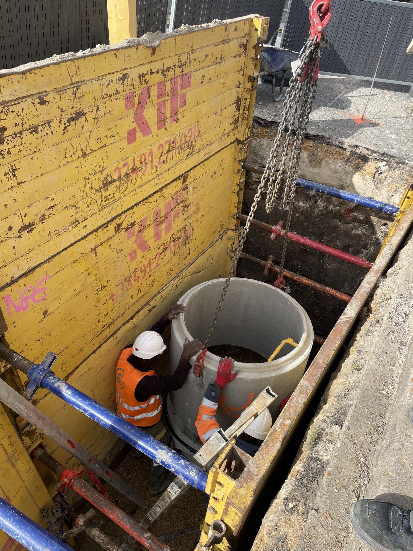 Des ouvriers du bâtiment installent un gros tuyau en béton dans une tranchée. Murs de protection jaunes, grue et équipements de sécurité.