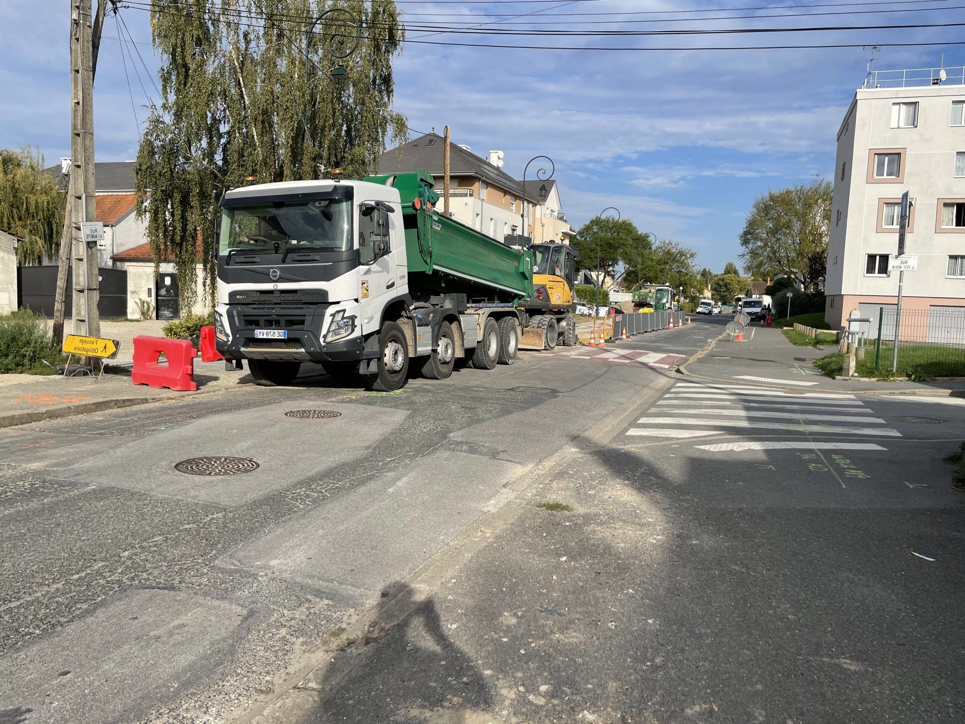 Un camion de chantier est stationné dans une rue en travaux ; un passage piéton est visible et des bâtiments bordent la rue.