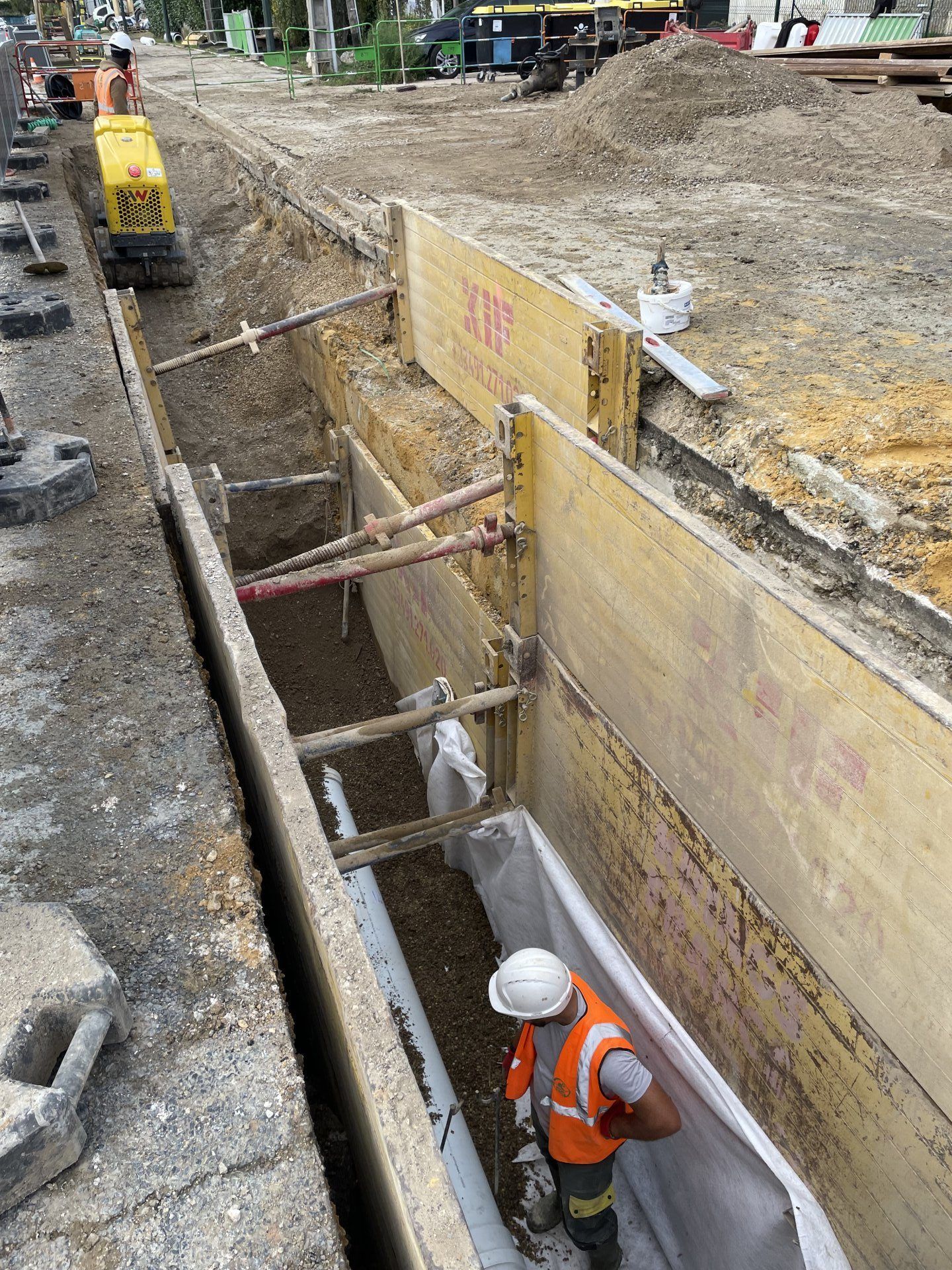 Un ouvrier du bâtiment travaille dans une tranchée revêtue de bois et d'étais, en bordure de route. On aperçoit une machine de compactage.