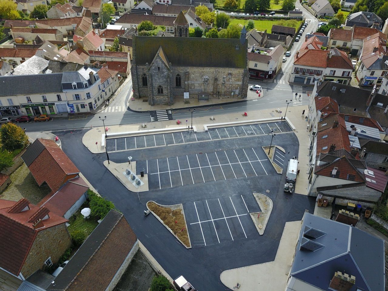 Vue aérienne d'une place de ville avec une église, un parking et des bâtiments environnants aux toits rouges.
