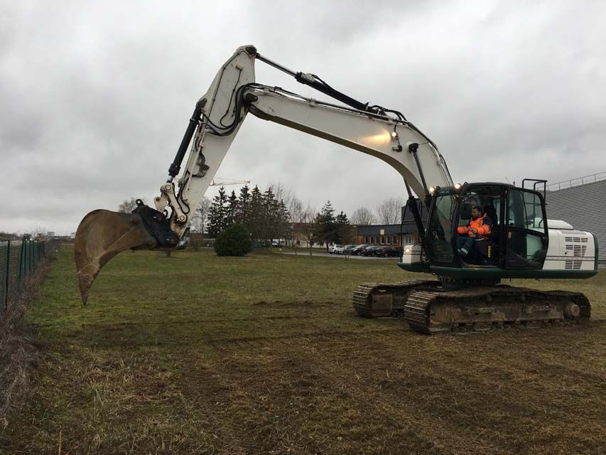 Une pelle mécanique blanche creuse un terrain herbeux. Un opérateur est dans la cabine, sous un ciel nuageux.