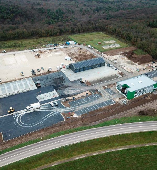 Vue aérienne d'un complexe comprenant plusieurs bâtiments, des travaux de construction, un parking et une route dans une zone boisée.