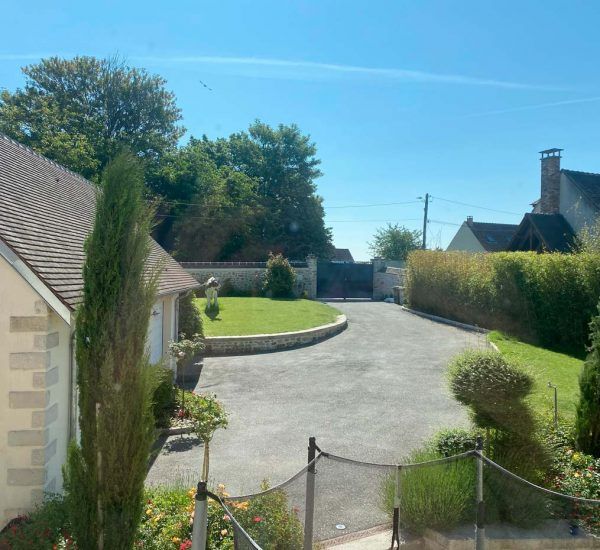 Allée menant à une propriété fermée par un portail, sous un ciel d'un bleu éclatant. Pelouse verdoyante et arbres entourent l'allée.